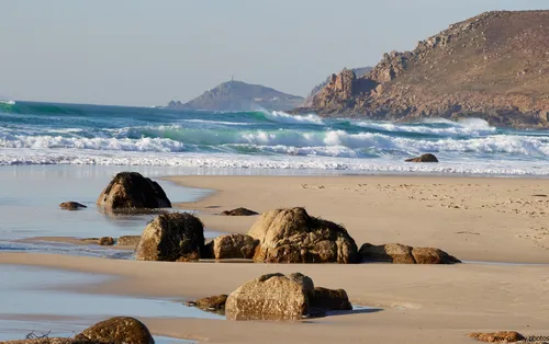 This landscape photograph shows Cape Cornwall viewed from Sennen beach on the Cornish coast in the United Kingdom during a spring afternoon. The scene captures the natural beauty of Sennen Cove, with waves breaking onto the sandy beach and several large rocks scattered across the shoreline. The rugged cliffs of Cornwall rise in the background, adding depth to the image and highlighting the region’s distinctive coastline. Cape Cornwall, a well-known landmark, is visible in the distance, with its promontory and historic structure clearly identifiable. The combination of coast, nature, cliffs, and beach under soft spring sunlight illustrates the character of Cornwall’s western shores.